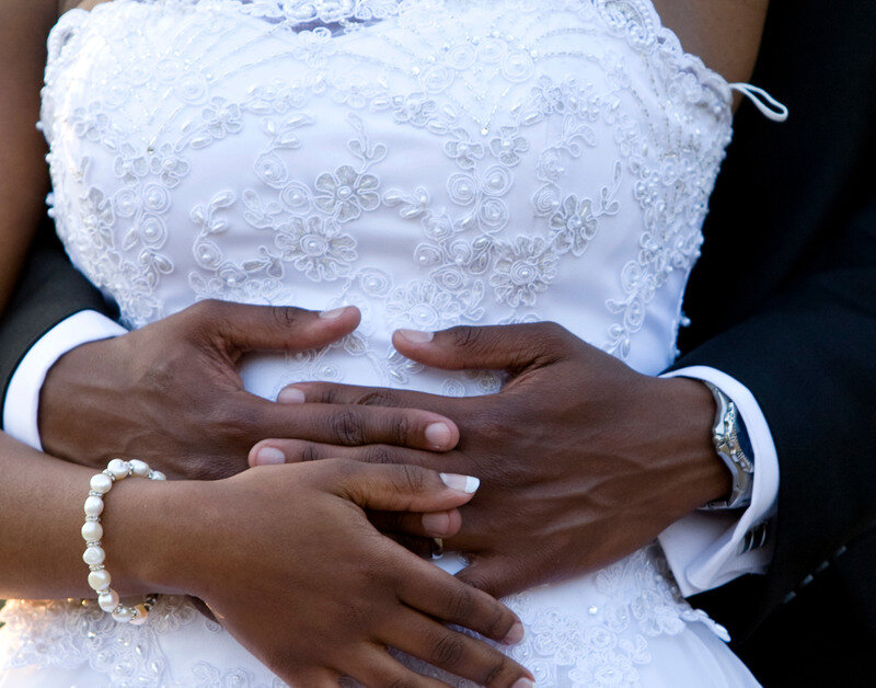A groom standing behind his bride, holding her by the waist. She rests her hands on his. They are outside at golden hour.