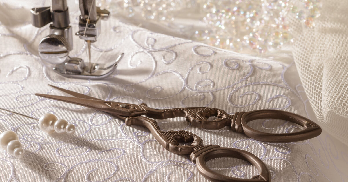 A sewing machine creating intricate patterns on a wedding dress. Scissors and pins rest on the material.