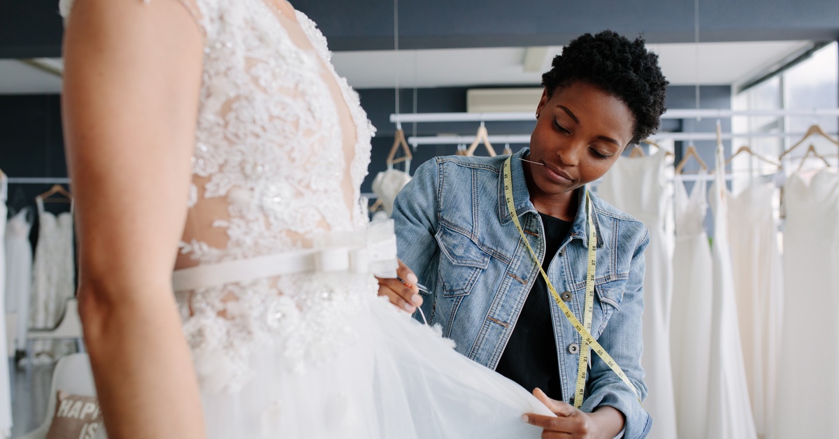 A professional designer holds onto a bride's gown to ensure proper fitting. She uses a measuring tape for precision.
