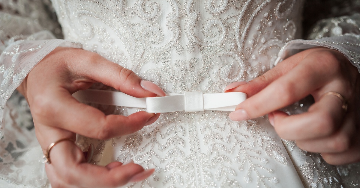 A waist-level view of a bride fastening a delicate bow on an intricately embellished wedding dress that shines.