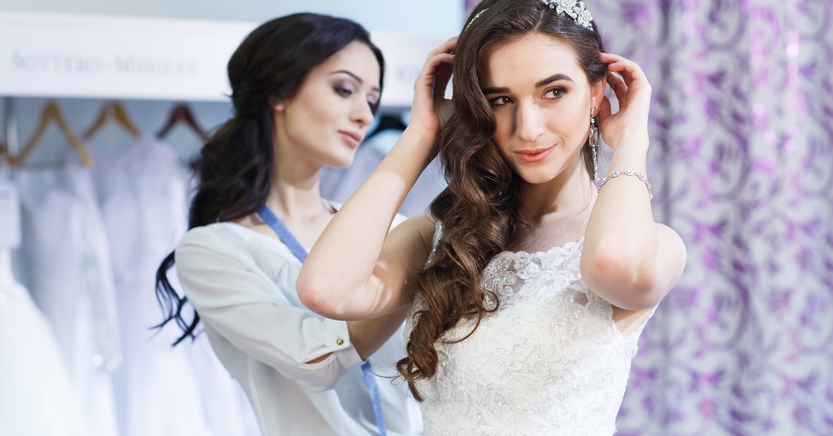 A bride smiles as she tries on a white dress at a shop. A professional tailor adjusts the back of her gown.