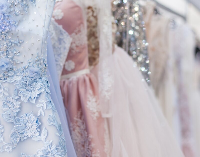 A rack of elegant evening dresses in blue, pink, and beige colors. The one in front is blue with floral embroidery.