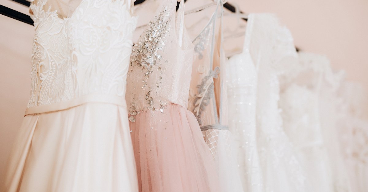 Several wedding gowns hang from a rack in a room. They have elegant designs with embroidery and vary in color.