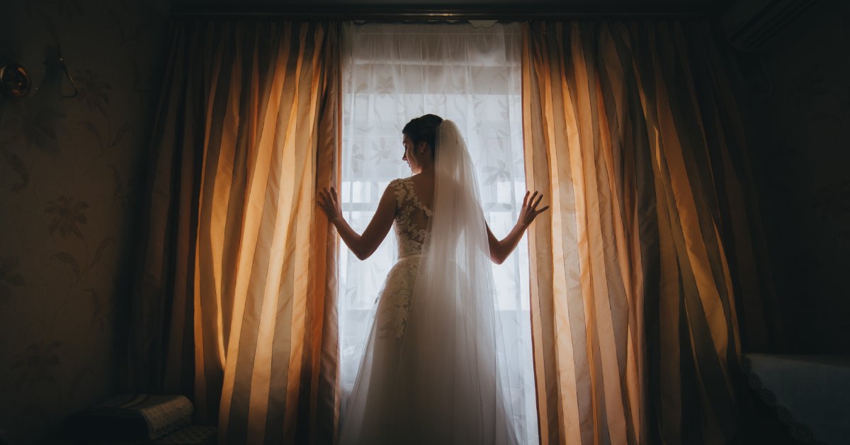 A bride wears a long veil and a white dress. She stands between two curtains by a window, creating a silhouette.