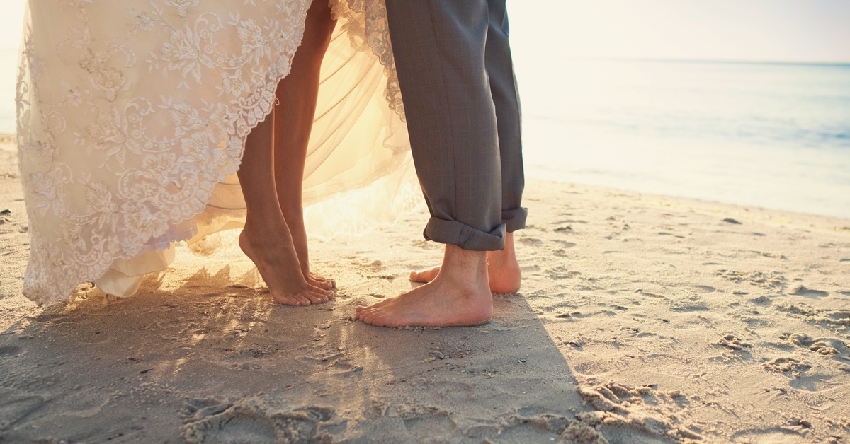 A bride and groom from the waist down standing barefoot on a sandy beach. The bride is on her tiptoes to kiss the groom.