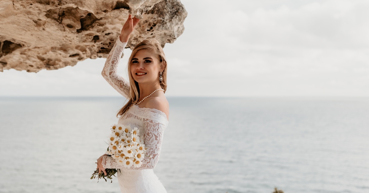 A bride poses by a rock formation near an ocean shoreline. She holds a daisy bouquet and wears a pearl necklace.