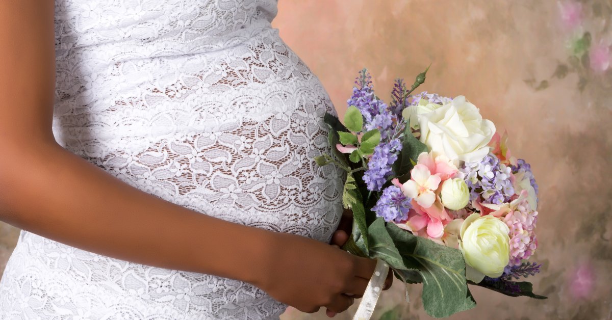 A woman dressed in a lacy wedding gown holds a small, beautiful bouquet in front of her pregnant belly.