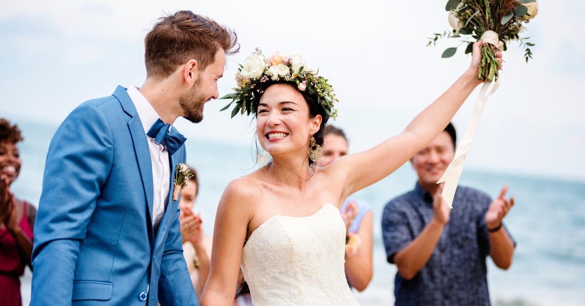 A bride, wearing a strapless dress, holds a bouquet up and smiles at the groom as a crowd applauds behind them.