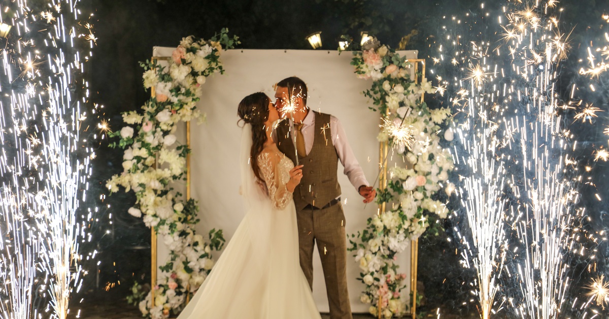 A wedding couple kisses in front of a floral background, surrounded by sparklers during a nighttime celebration.